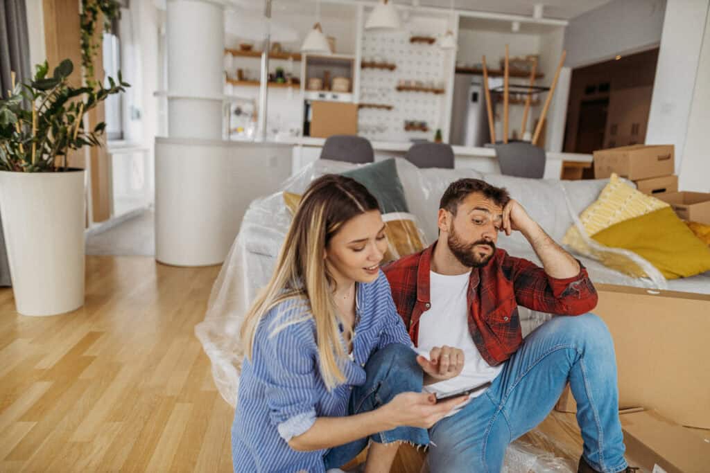 Jong stel zit tussen verhuisdozen in hun nieuwe woning en bekijkt samen iets op een smartphone. De man oogt vermoeid en de vrouw probeert iets uit te leggen, passend bij de keuzestress tussen huren en kopen op de krappe woningmarkt van 2025.