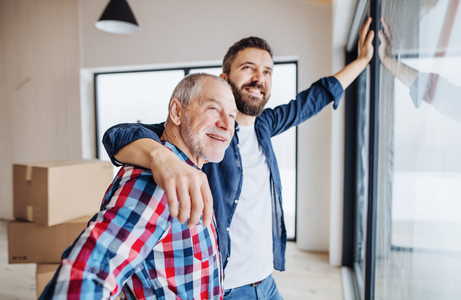 Volwassen zoon en vader kijken samen uit het raam van een nieuw huis – een symbolisch moment van familiehulp bij het kopen van een woning.