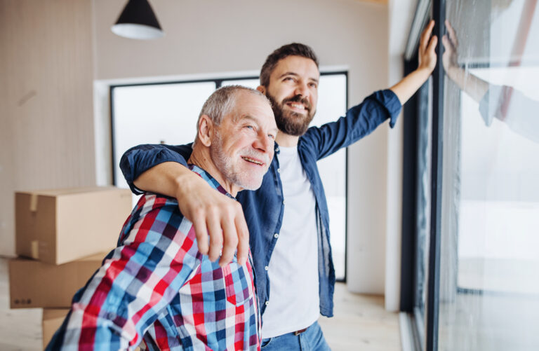 Volwassen zoon en vader kijken samen uit het raam van een nieuw huis – een symbolisch moment van familiehulp bij het kopen van een woning.
