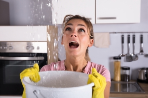 Vrouw in een keuken, met een angstige uitdrukking, houdt een emmer onder een lekkage die water van het plafond opvangt. Ze draagt rubberen handschoenen terwijl het water van bovenaf naar beneden stroomt