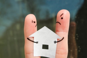 Twee vingers met daarop getekende gezichten - één vrolijk en één boos - houden een papieren huisje vast, wat symbool staat voor de uitdagingen rondom de verdeling van een huis bij een scheiding.