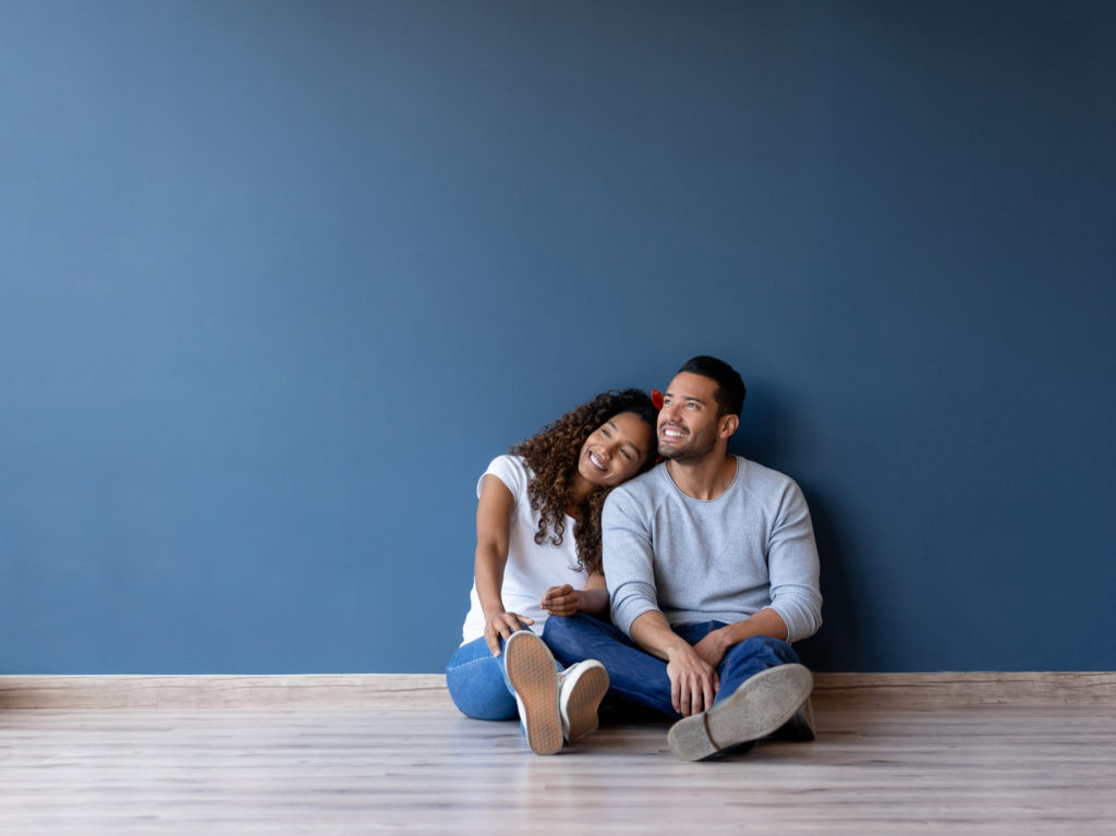 Een lachend stel zit ontspannen op de vloer tegen een blauwe muur in een pas gerenoveerde ruimte, genietend van een warm en comfortabel huis.