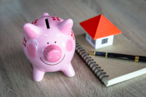 Een spaarvarken, notitieboekje en miniatuurhuisje op een houten tafel, symbool voor het plannen en besparen van energie in huis.