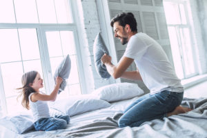 Vader en dochter springen samen op een boxspring bed in een lichte, moderne slaapkamer met witte bakstenen muren en grote ramen