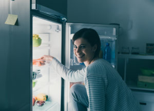 Vrouw glimlacht terwijl ze iets uit de koelkast pakt, een huishoudelijk apparaat gevuld met vers voedsel. Er hangt een briefje met een hartje op de koelkastdeur