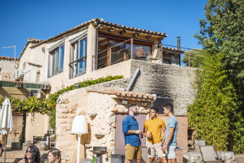 Drie mannen genieten van een gesprek buiten bij een rustiek huis in Zuid-Europa, met de charme van traditionele architectuur en zonnig weer.