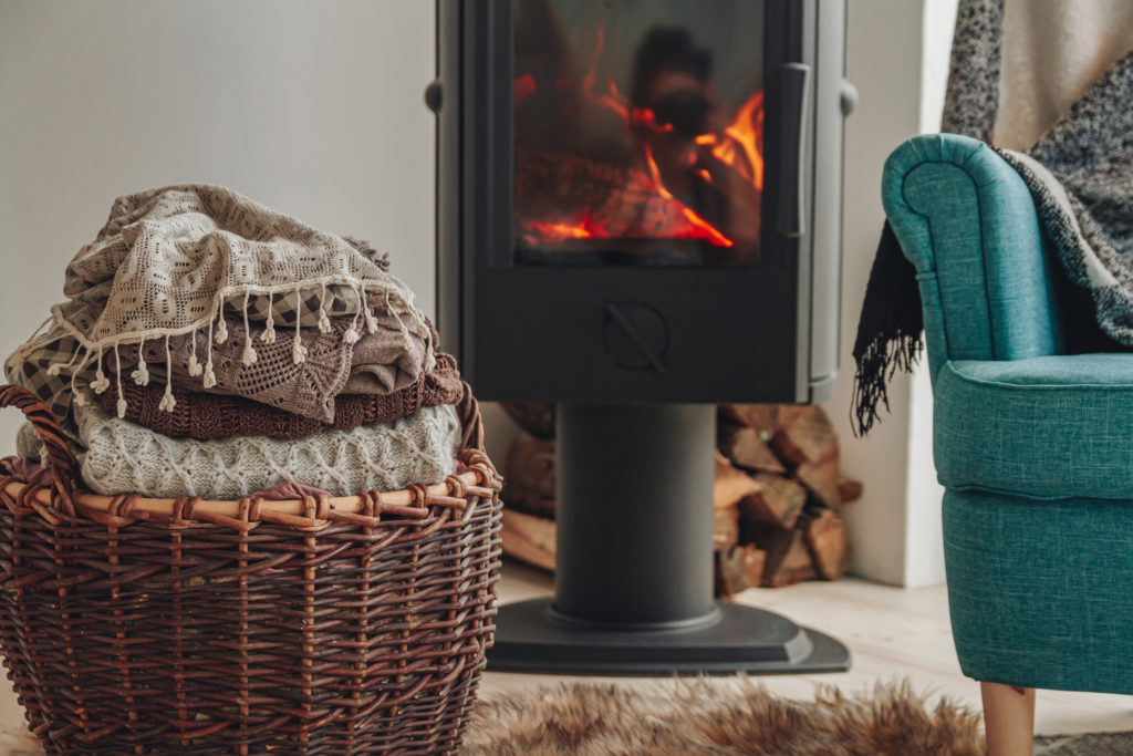 Gezellige woonkamer met een brandende houtkachel, een mand vol zachte dekens, en een comfortabele blauwe stoel, met brandhout opgestapeld naast de haard.