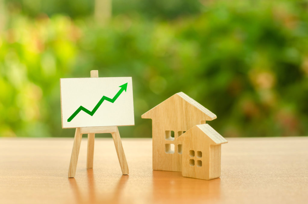 Twee houten huisjes met een bord waarop een stijgende groene lijn is getekend, wat suggereert dat de waarde van de huizen toeneemt.