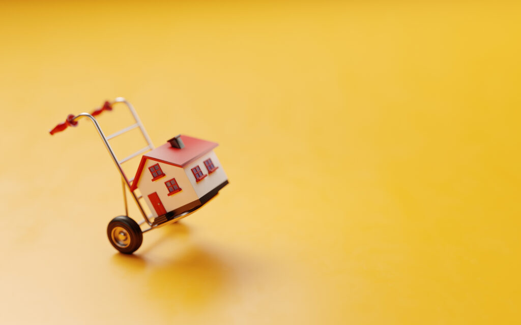 Miniatuurhuisje op een steekwagen tegen een gele achtergrond, symbool voor verhuizing. Het beeld benadrukt het concept van het verplaatsen van een woning, ideaal voor gebruik in contexten van verhuisdiensten.