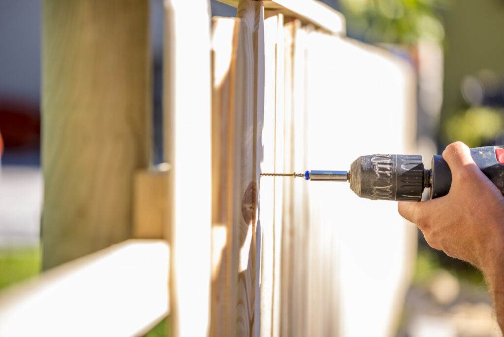 Man gebruikt een elektrische boormachine om een houten schutting te monteren in een zonnige tuin
