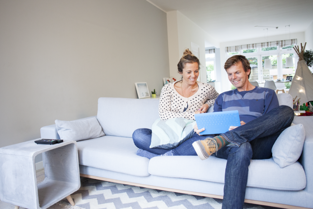 Een glimlachend stel dat op de bank samen naar een tablet kijkt, symboliserend het belang van woon- en inboedelverzekeringen voor gemoedsrust.