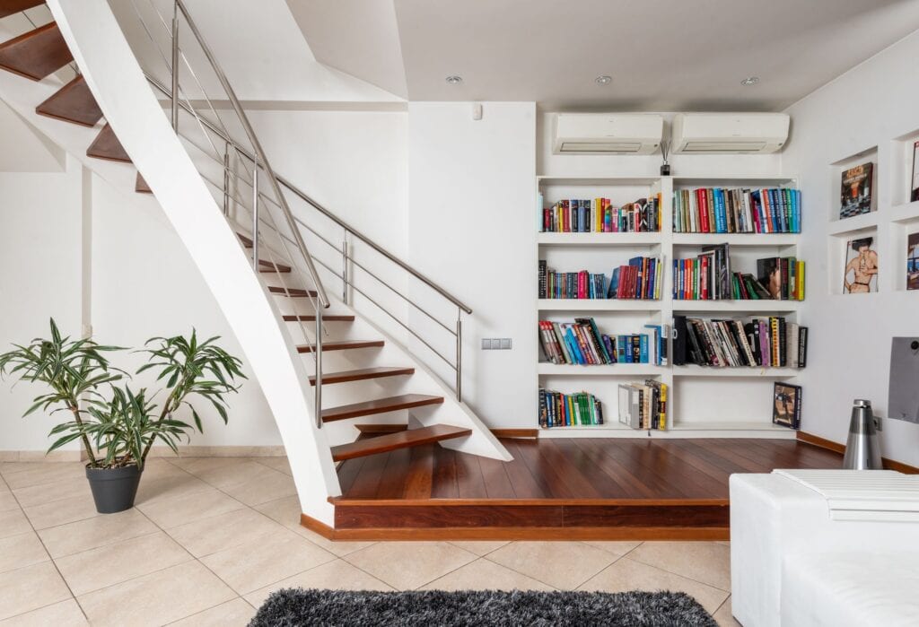 Moderne woonkamer met zwevende houten trap, metalen trapleuning, ingebouwde boekenkast en kamerplant in pot. De kamer heeft een strakke, minimalistische uitstraling met lichte muren, tegelvloer en houten vloerdelen.
