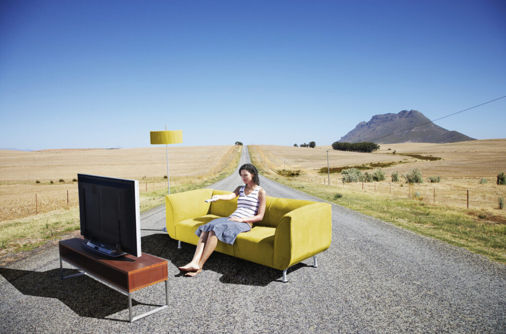 Een vrouw zit op een gele bank midden op een lange, lege weg, met een TV en lamp naast haar – een surrealistisch beeld dat de balans tussen inrichting en functionaliteit benadrukt.