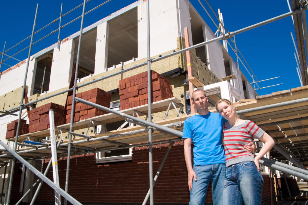 Een jong stel poseert voor hun nieuwbouwwoning in aanbouw, omringd door steigers en bouwmaterialen, symbool voor het proces van het bouwen van een nieuwbouwwoning.