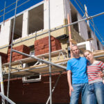 Een jong stel poseert voor hun nieuwbouwwoning in aanbouw, omringd door steigers en bouwmaterialen, symbool voor het proces van het bouwen van een nieuwbouwwoning.