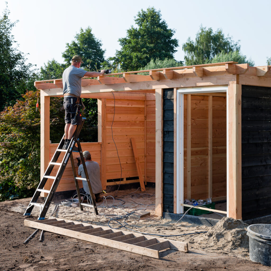 Twee mannen werken aan de bouw van een houten tuinhuisje, waarbij één op een ladder staat om het dak te bevestigen, terwijl de ander binnenin bezig is met boren.