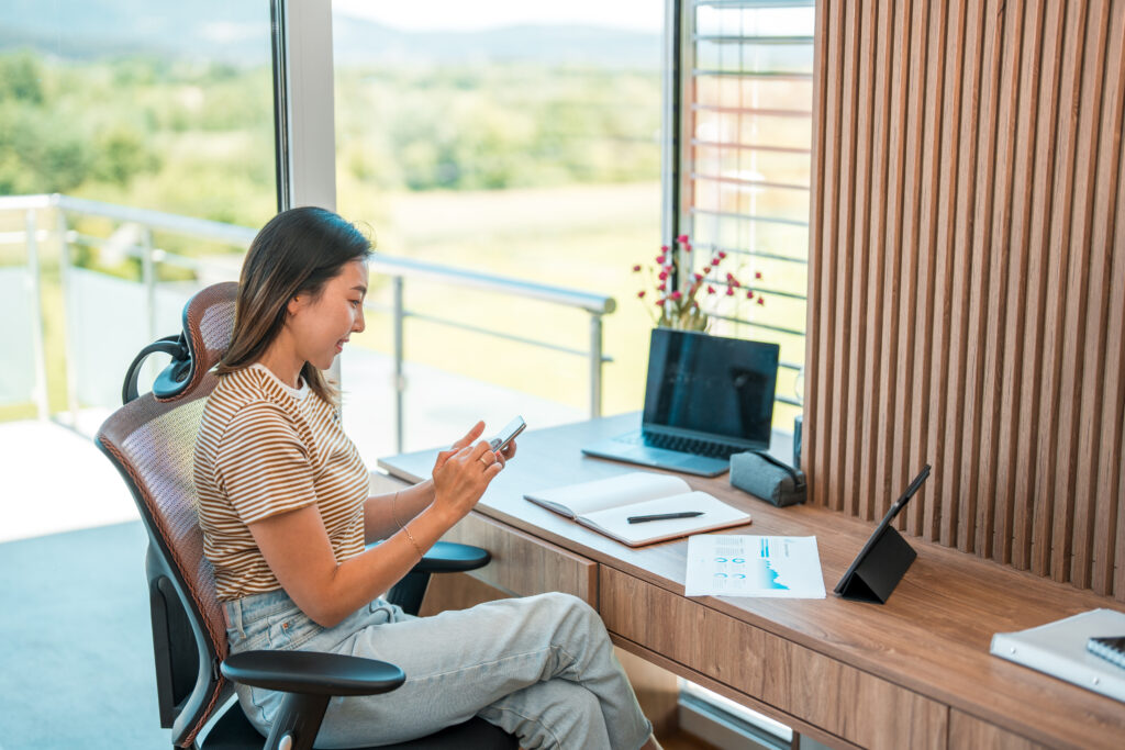 Een vrouw zit in een moderne en lichte thuiswerkomgeving, werkend aan een houten bureau met een laptop, tablet en documenten. Op de achtergrond is een groene buitenomgeving zichtbaar door grote ramen.