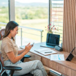 Groothoekopname van een vrouw in vrijetijdskleding die typt op een smartphone, haar bureau vol met aantekeningen en accessoires, een laptop en een digitale tablet.