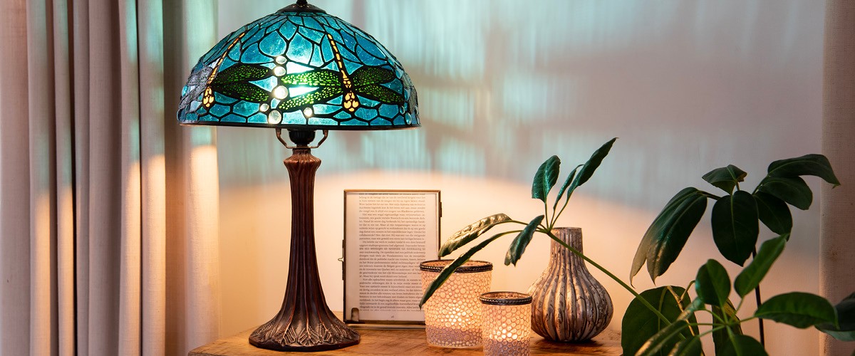 Een sfeervol verlichte Tiffany-lamp met een gekleurd glazen kap, versierd met libellen, staat op een houten tafel naast enkele decoratieve voorwerpen en planten