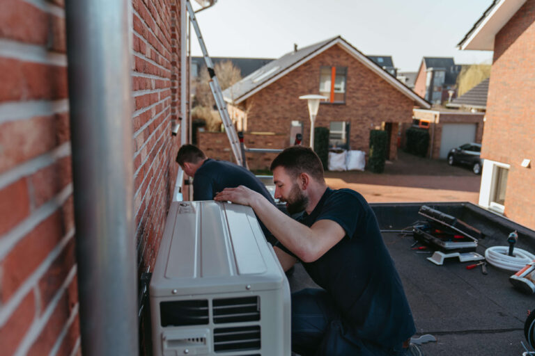 Twee monteurs installeren een split airco-unit aan de buitenmuur van een woning, met gereedschap op de grond in een residentiële omgeving.