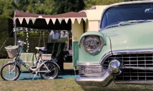 Retro stacaravan op een camping met een vintage auto en een fiets op de voorgrond, ingericht als recreatiewoning met een luifel en decoratieve vlaggen.