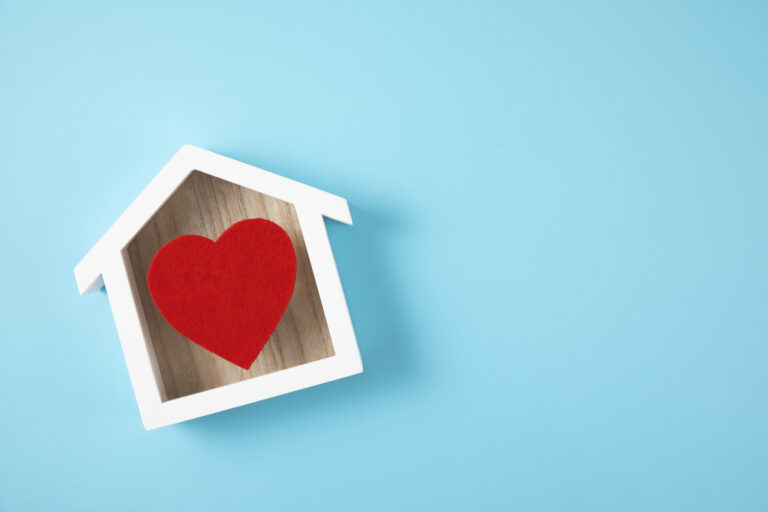 Houten huisvorm met een rood hart in het midden tegen een lichtblauwe achtergrond. Perfect symbool voor het creëren van een warm en samenhangend interieur na het kopen van een huis.