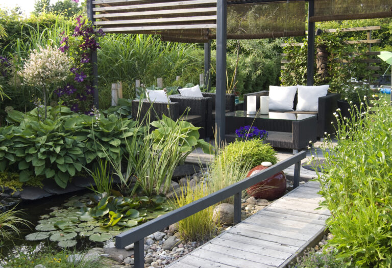 Moderne tuin met donkere accenten: stijlvolle loungehoek met zwarte rotanmeubels onder een pergola, omringd door weelderige planten en een vijver vol lelies. Donkere accenten voegen contrast toe aan het groene landschap en creëren een rustgevende sfeer