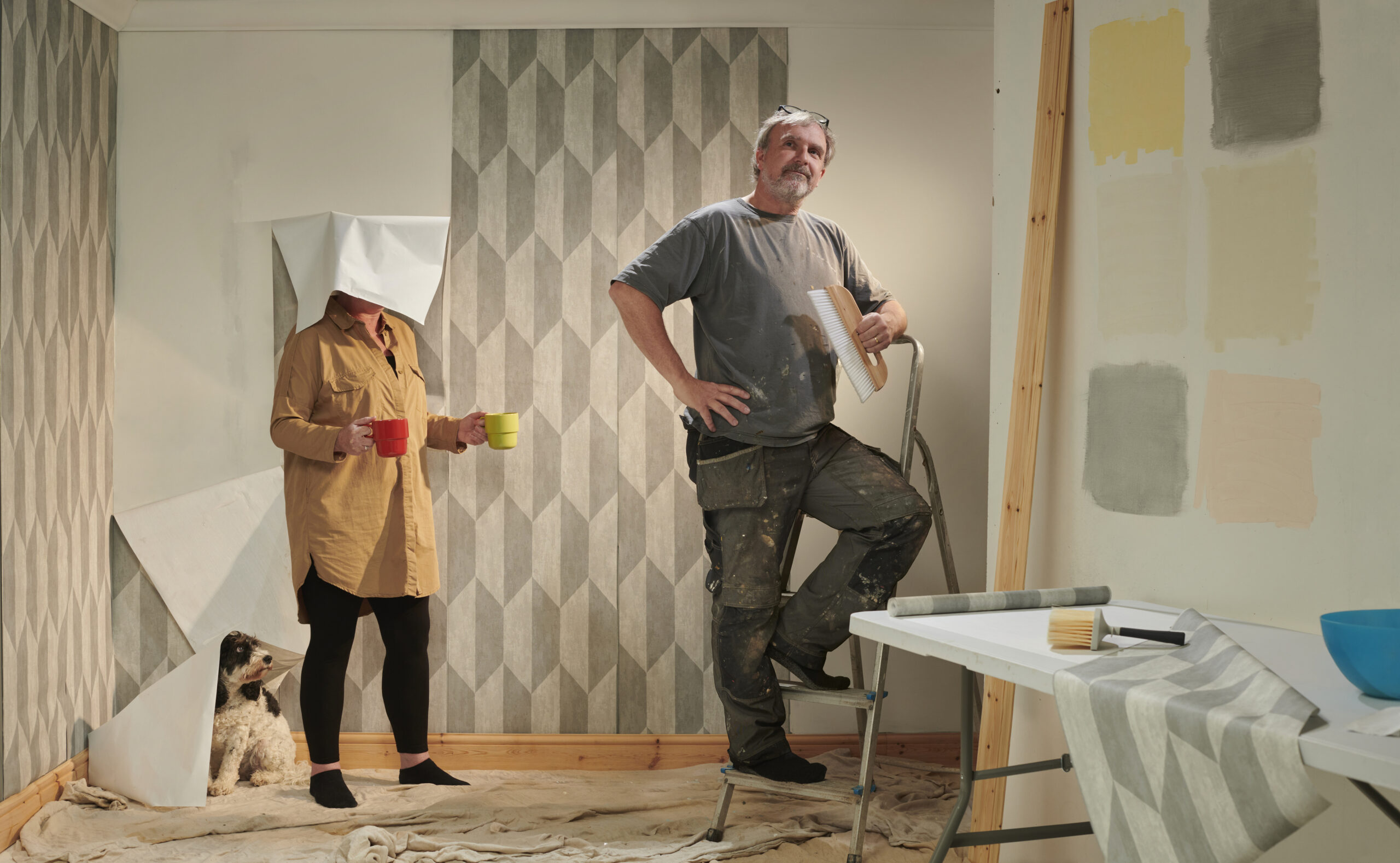 Behangen van nieuwbouwwoningen: Een stel tijdens het behangen met patroonbehang, verfkleuren op de muur en een nieuwsgierige hond in de kamer.