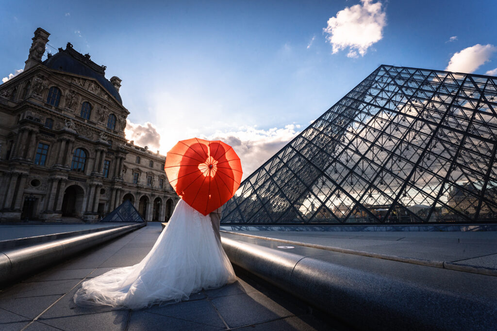 ruidspaar met hartvormige rode paraplu poseert voor de glazen piramide van het Louvre bij zonsondergang, ideaal voor een XL wanddecoratie.