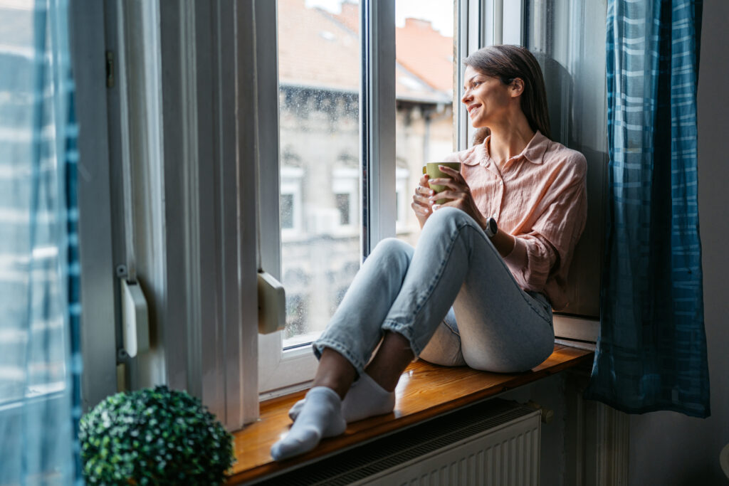 Vrouw zit ontspannen bij een raam met een kopje in de hand, genietend van een comfortabele en seizoensgebonden sfeer in huis.