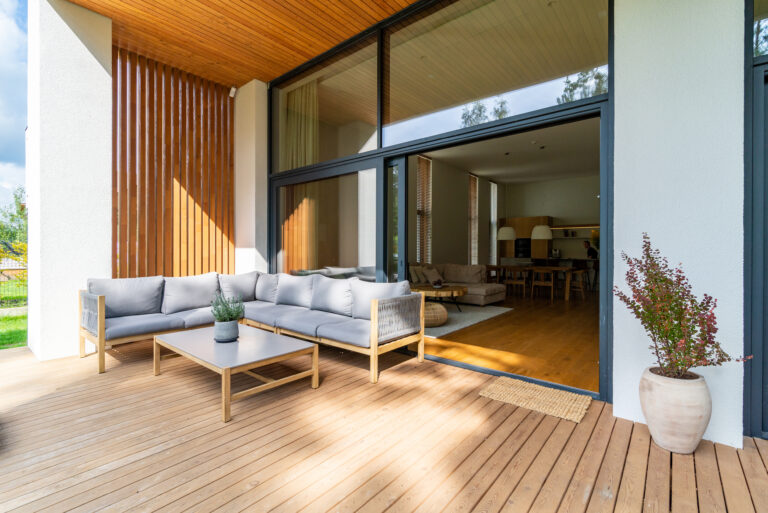 Een stijlvolle houten veranda met een ruime loungebank, grenzend aan een moderne open woonkamer met grote glazen schuifdeuren, benadrukt de naadloze overgang tussen huis en tuin.