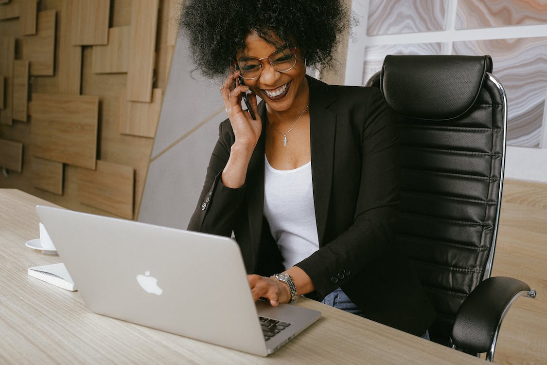 Zakelijke vrouw in een ergonomische bureaustoel, lachend terwijl ze werkt op een laptop en belt in een moderne kantooromgeving