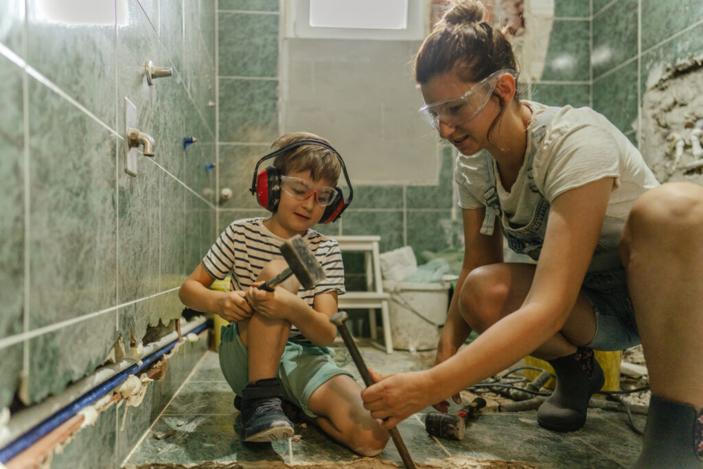 Een moeder en haar zoon dragen veiligheidsbrillen terwijl ze samen in een badkamer werken tijdens een verbouwing, omringd door gereedschap en verbouwingsmaterialen.