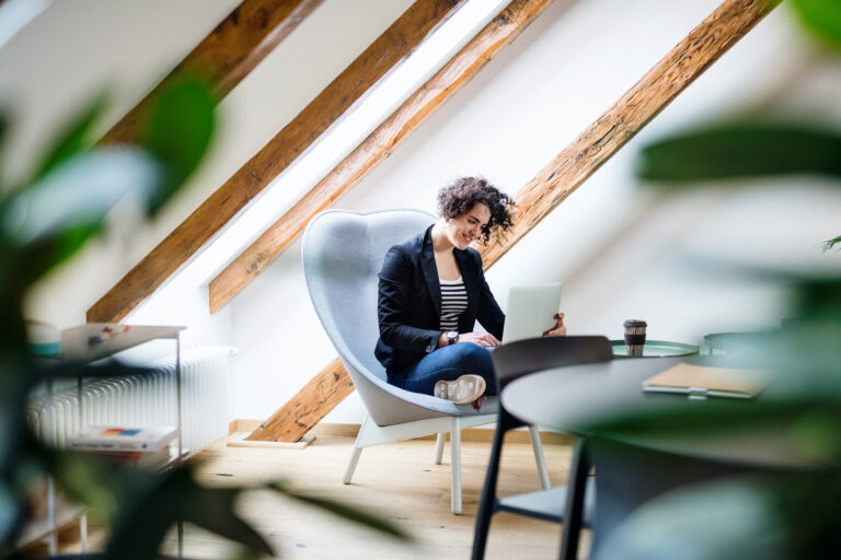 Een persoon werkt ontspannen met een laptop in een modern thuiskantoor op zolder, omgeven door sfeervolle houten balken en veel natuurlijk licht. Perfect voorbeeld van een slim en energiezuinig ingerichte werkruimte