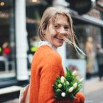 Blonde vrouw in een oranje trui loopt lachend door een winkelstraat met een bos witte tulpen in haar hand.