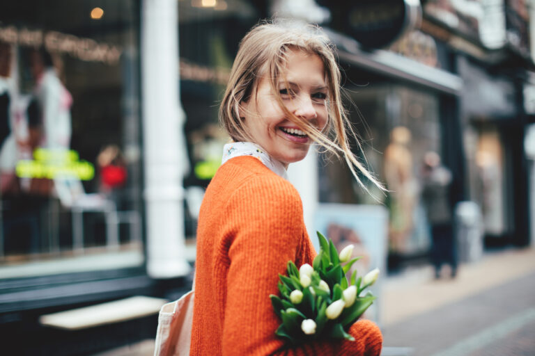 Blonde vrouw in een oranje trui loopt lachend door een winkelstraat met een bos witte tulpen in haar hand.