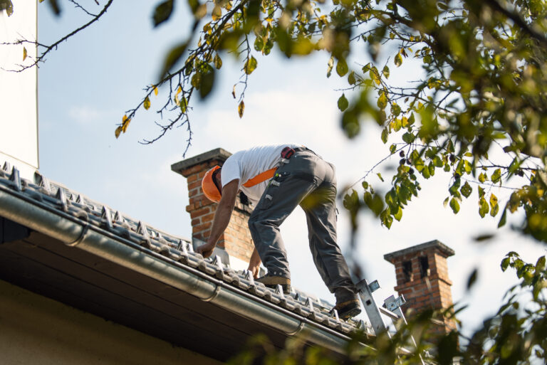 Dakdekker op een ladder voert inspectie en reparatie uit aan een dak, met zicht op bakstenen schoorstenen en omliggende bladeren.