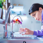 Vrouw kijkt bezorgd naar een kraan met lage waterdruk in een keuken, terwijl ze een glas vasthoudt.