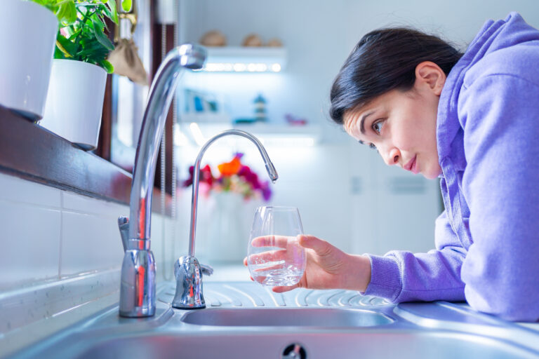 Vrouw kijkt bezorgd naar een kraan met lage waterdruk in een keuken, terwijl ze een glas vasthoudt.