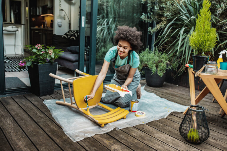 Vrouw die houten tuinmeubelen schildert in een groene tuin, met verf en kwast in de hand.