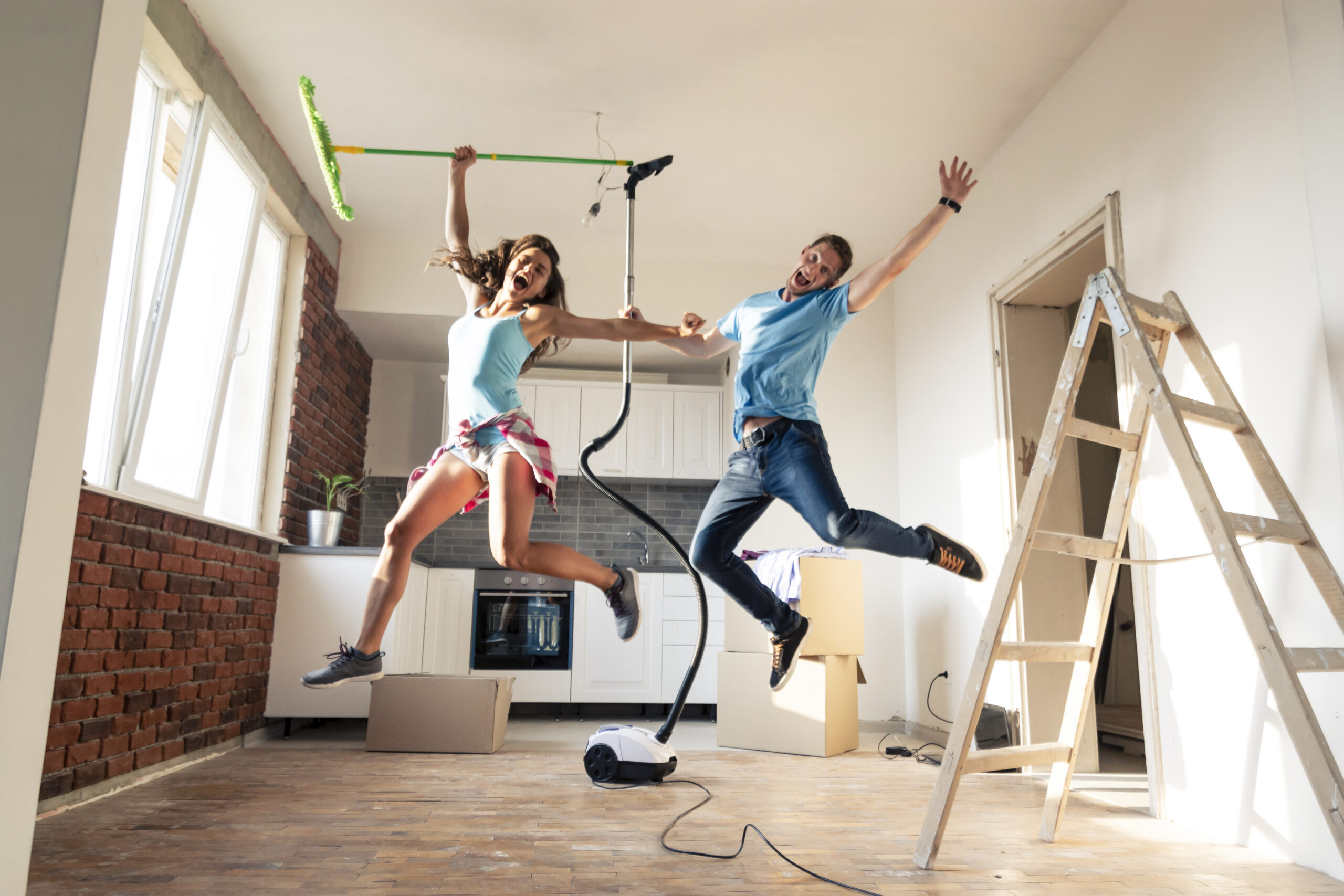 Een vrolijk stel springt in de lucht terwijl ze schoonmaken met een stofzuiger in een lichte, moderne woonkamer. De afbeelding straalt energie en plezier uit tijdens het schoonmaken.