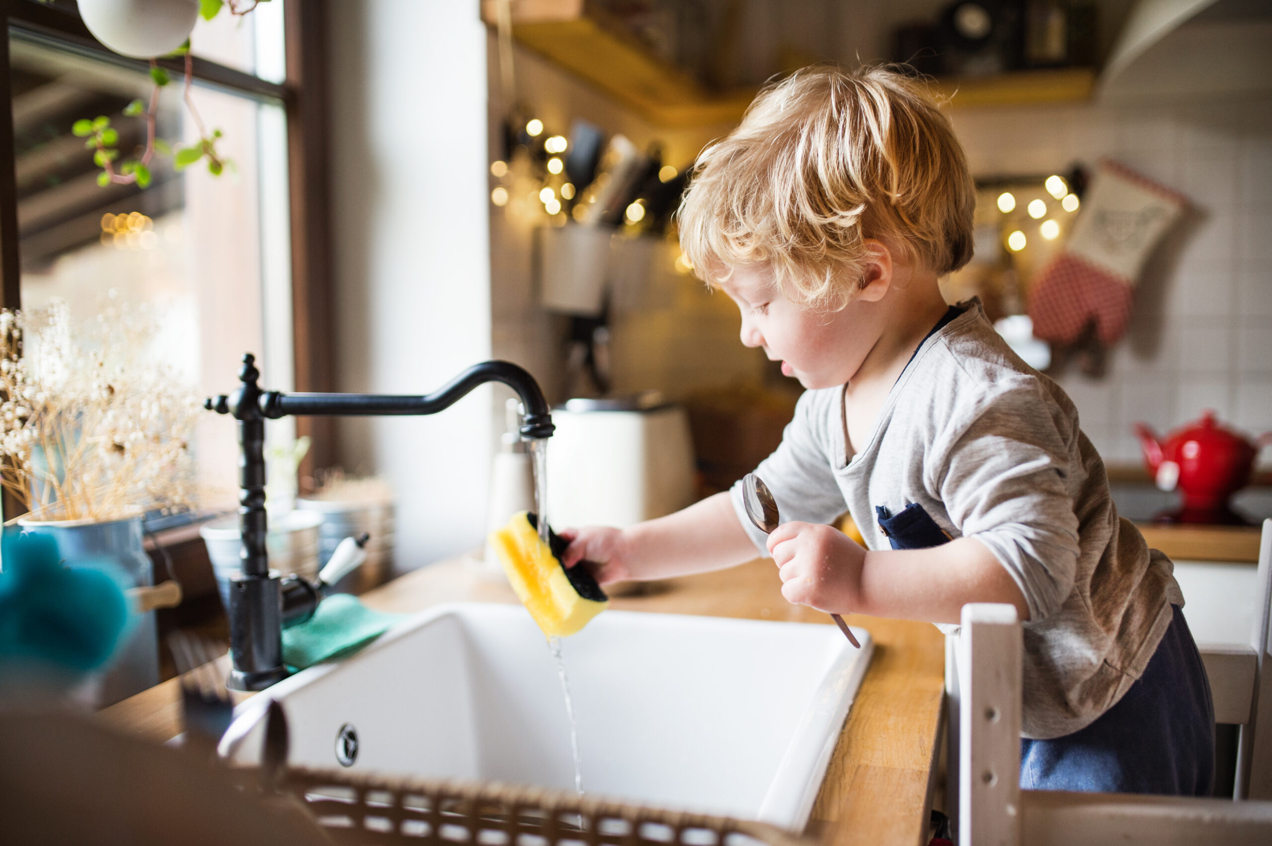 Klein jongetje wast een lepel bij de gootsteen in een keuken, met stromend water uit de kraan en een spons, symboliserend het gebruik van zacht water dankzij een waterontharder.