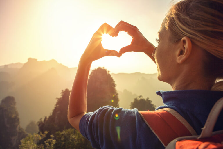 Vrouw maakt hartje met handen in berglandschap – symbool voor ontspannen reizen tijdens verbouwing.