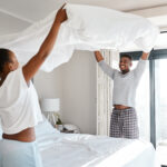 Koppel maakt samen het bed op met fris wit beddengoed in een lichte, sfeervolle slaapkamer met veel daglicht.
