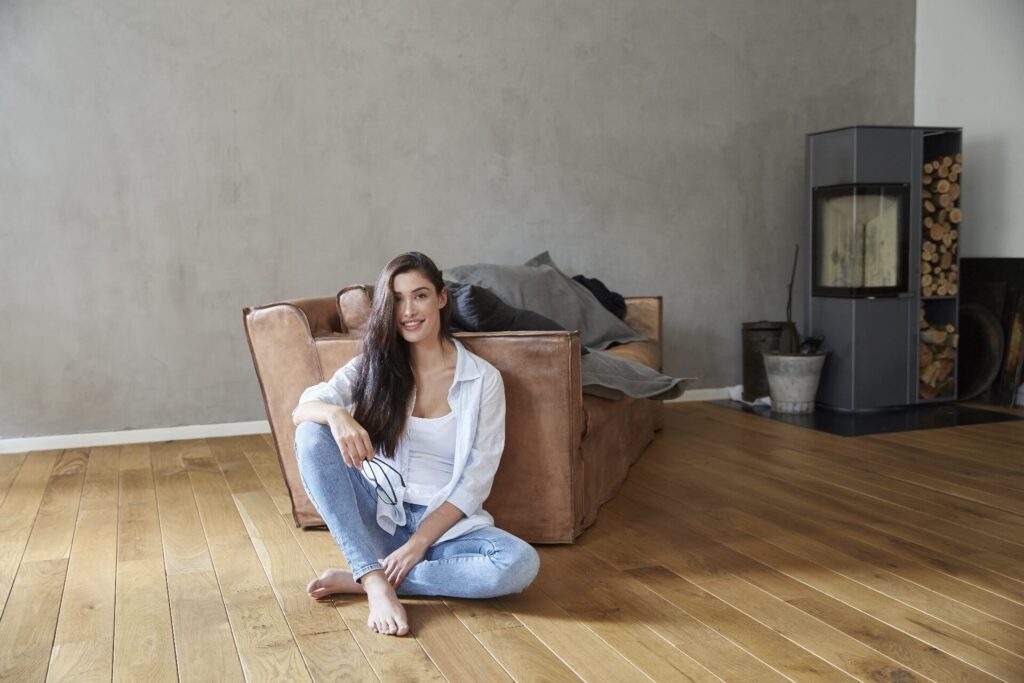 Vrouw zit ontspannen op een houten vloer met brede planken in een moderne woonkamer met leren bank en houtkache