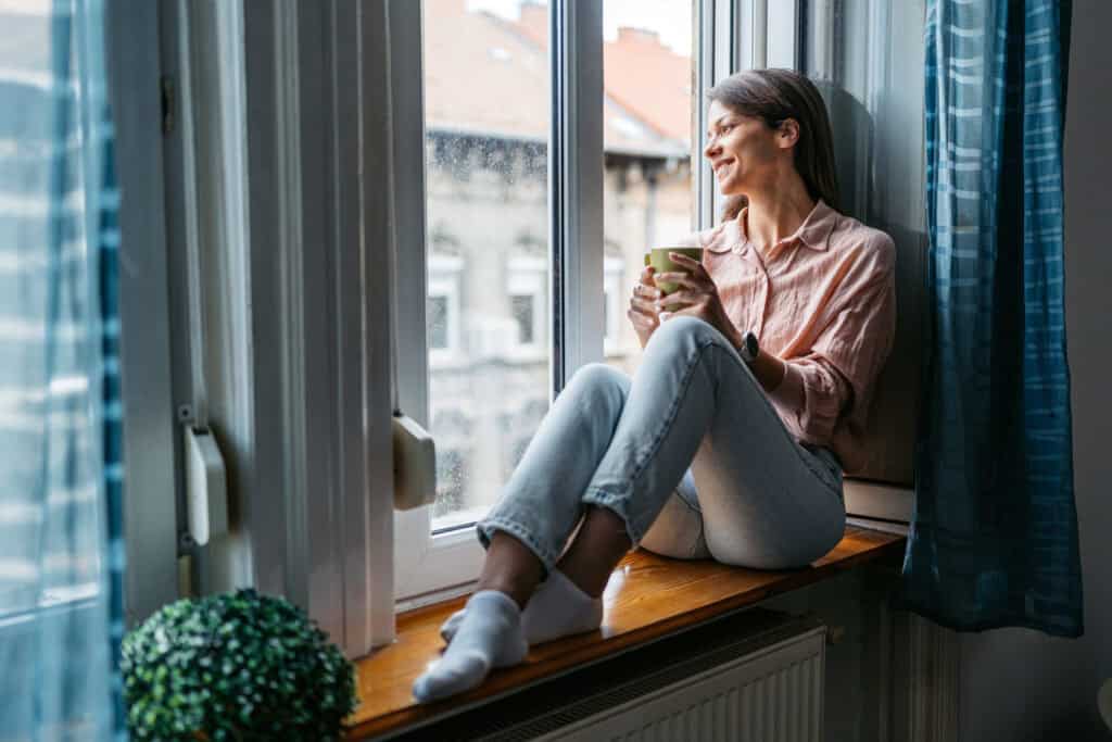 Vrouw met kopje thee zit op vensterbank bij groot raam, comfortabel en warm