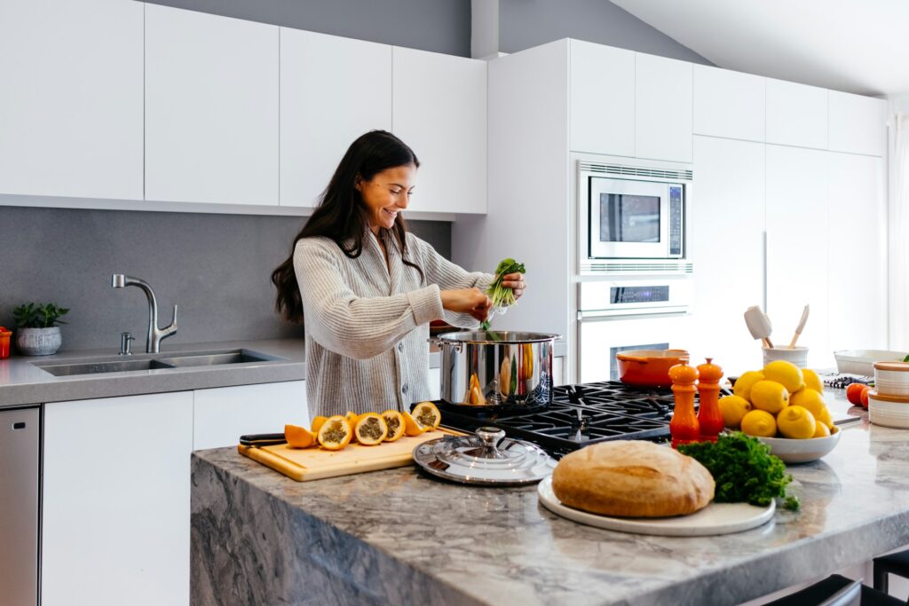 Vrouw kookt met verse groenten in een moderne, lichte keuken met RVS-pan, brood en citrus op het aanrecht