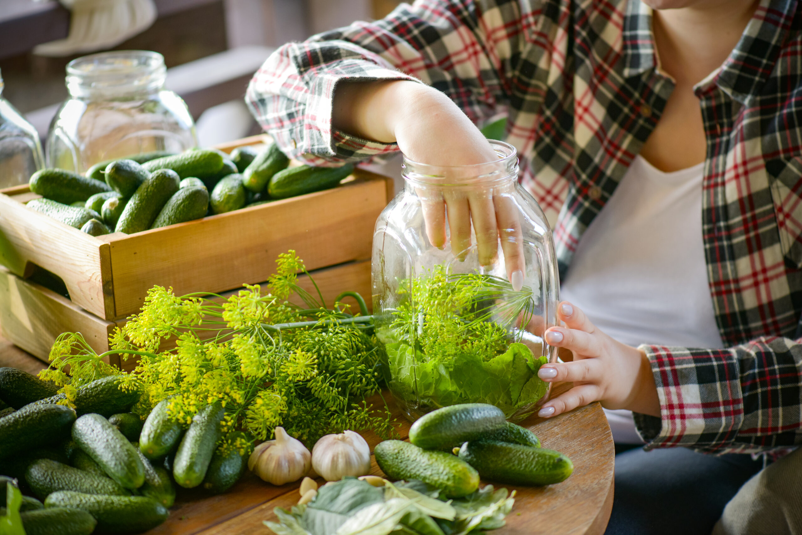 Persoon bereidt verse kruiden en komkommers in glazen potten aan een houten keukentafel, omringd door natuurlijke ingrediënten zoals knoflook en dille