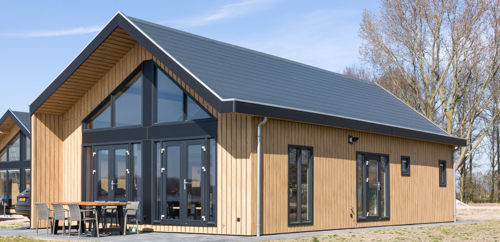 Licht houten chalet met zwarte kozijnen, hoge ramen en terras onder een strak zadeldak