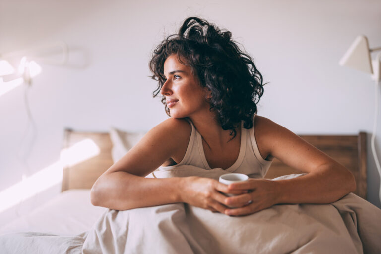 Vrouw zit ontspannen rechtop in bed met een kopje in haar handen, omringd door zacht ochtendlicht